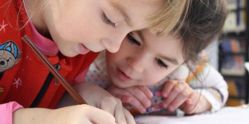 Zwei Mädchen im Grundschulalter beugen sich gemeinsam über ein Heft und Schreiben.