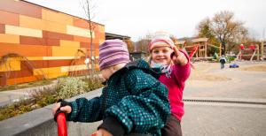 Nahaufnahme von zwei Kindern auf dem Dreirad, im Hintergrund der Spielplatz mit Sandkästen und Klettergerüsten