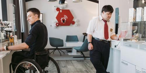 Ein junger Mann mit weißen Chucks und scharzer Kleidung im Rollstuhl sitzend und ein junger Mann mit Anzughose, weißem Hemd und roter Krawatte mit Down Syndrom arbeiten in einem modernen Café.