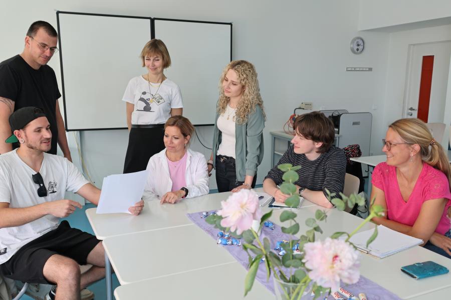 Sieben Erwachsene sitzen in einem Klassenraum um Tische herum